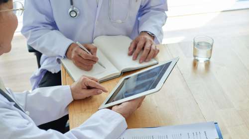 Group of experienced physicians analyzing results of chest X-ray with help of digital tablet, one of them taking necessary notes